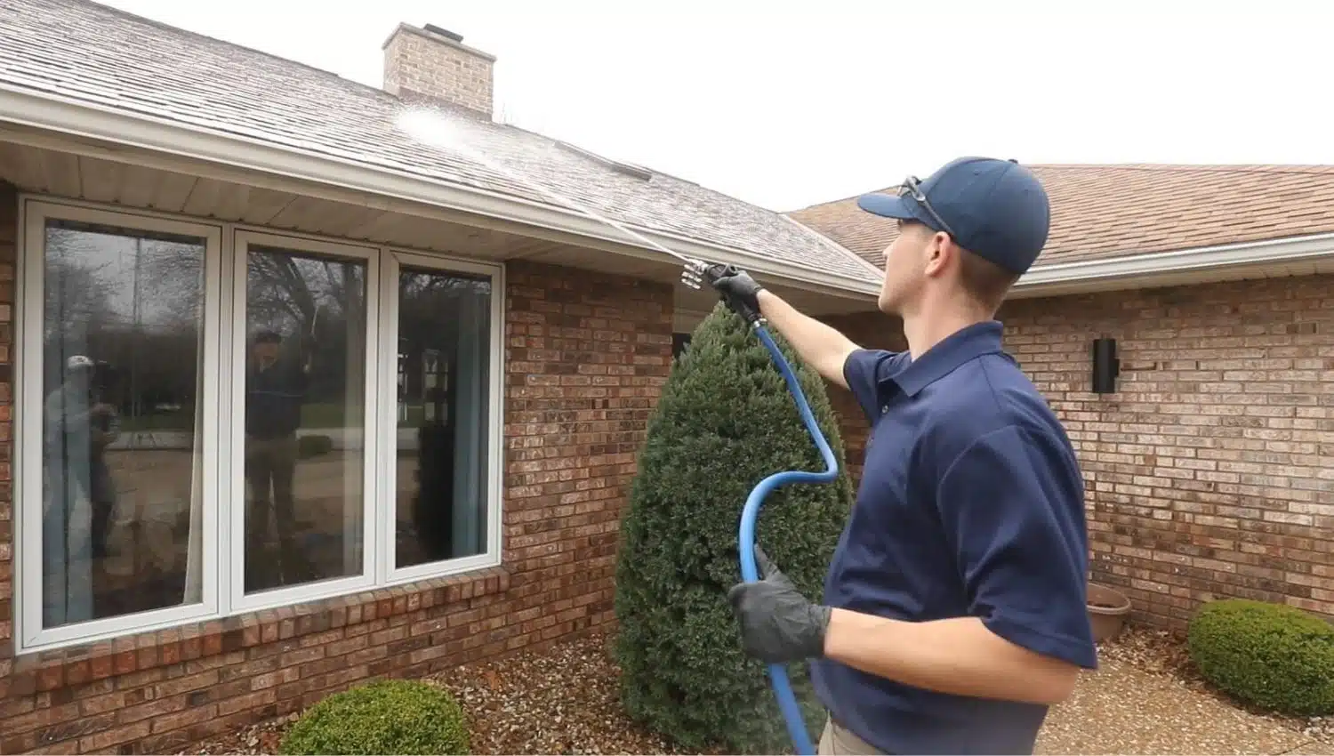 Roof Cleaning A man pressure washing a roof for roof cleaning. He wears a blue shirt, cap, and gloves, holding a blue hose, standing in front of a brick house with large windows and a trimmed shrub.